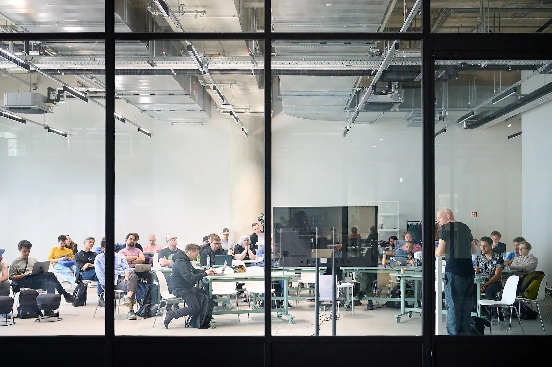 Our PouchDB workshop took place in a large open lecture room with industrial styling. In this image we’re peeking through the large glass windows into the workshop from the communal area, and see people gathered with laptops, following Alex’ demo. Photo by Susanne Elsässer.