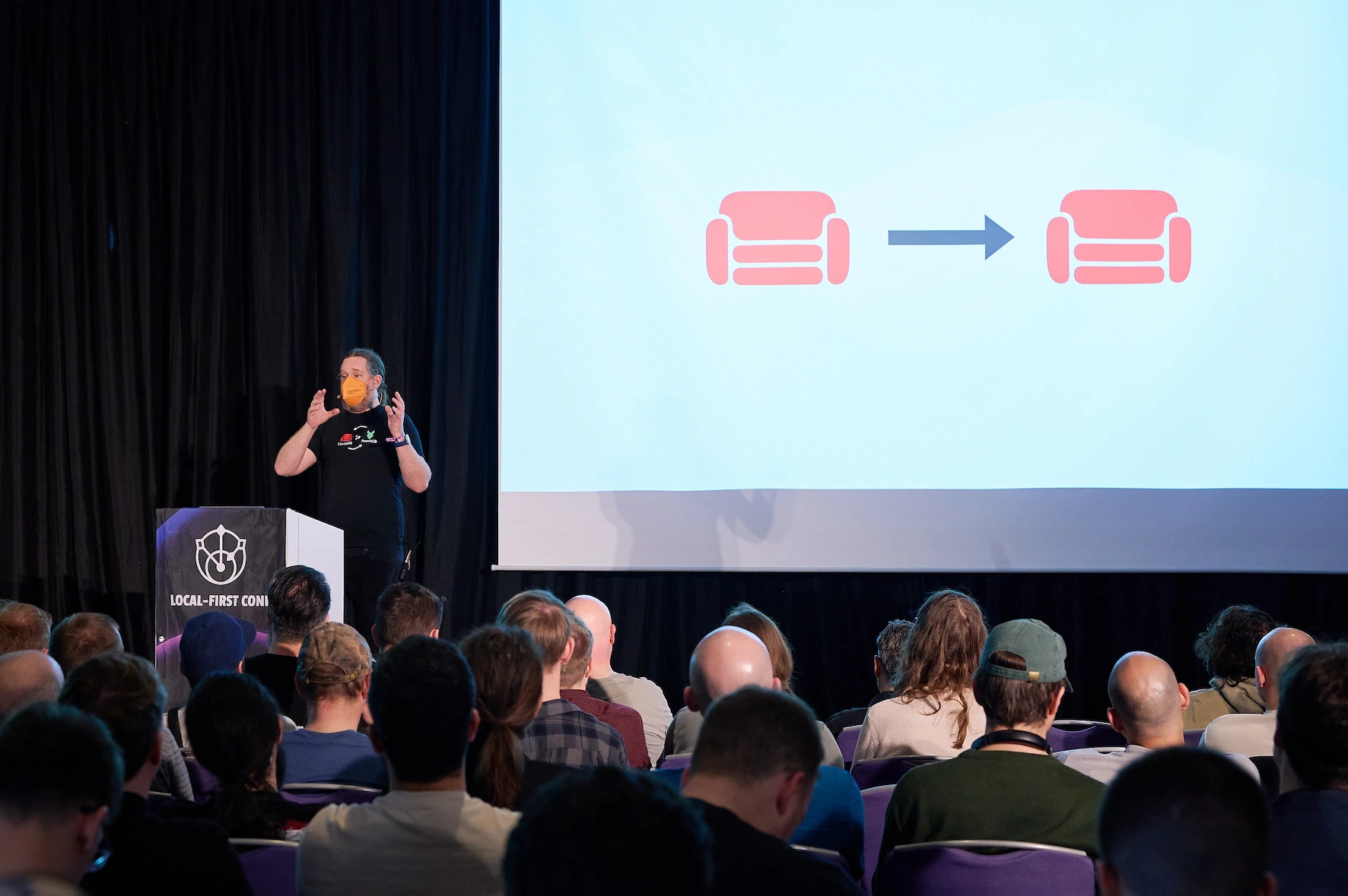 Jan Lehnardt stands in front of the Local First podium on stage at the conference. Behind him we two red sofas are projected, representing two CouchDB instances in sync. The conference is well attended: a large and engaged audience are taking notes.